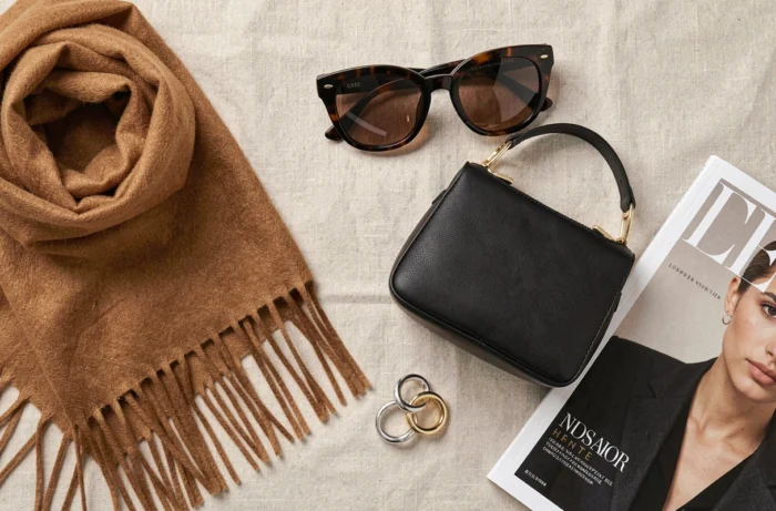 Flat-lay d'accessoires de mode tendance sur lin beige : écharpe cachemire, sac en cuir noir, lunettes et bijoux dorés